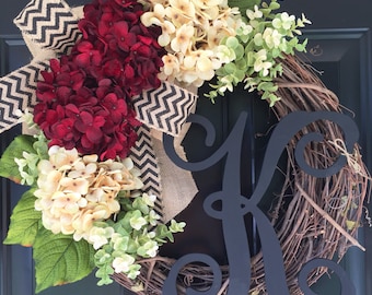 May include: A wreath made of grapevine with a black letter "K" in the center. The wreath is decorated with burgundy and cream hydrangeas, green leaves, and a black and white chevron ribbon.