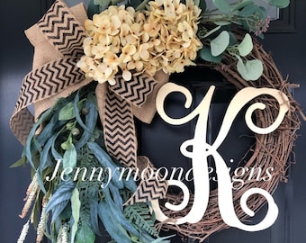 May include: A grapevine wreath adorned with faux beige hydrangeas, green foliage, and a burlap ribbon with a black chevron pattern. A large wooden letter "K" is centered on the wreath. The text "Jennymoon designs" is visible.