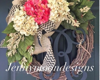 Può includere: Una ghirlanda di vite decorata con un fiocco di juta e un nastro a chevron bianco e nero. La ghirlanda è adornata con ortensie rosa e bianche e verde. Un monogramma "R" in metallo nero è posizionato al centro della ghirlanda. La ghirlanda è appesa a una porta nera. Il testo "Jennymoon Designs" è visibile nella parte inferiore dell'immagine.