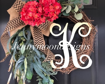 May include: A wreath with a burlap bow with black and white chevron pattern, red hydrangeas, greenery, and a white wooden letter K. The wreath is on a black door with the text "Jenny Moon Designs".