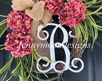 May include: A wreath with a burlap bow, pink hydrangea flowers, and green leaves. A white wooden letter "B" is in the center of the wreath. The wreath is on a black door. The text "Jennymoon Designs" is visible in the image.