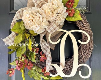 May include: A decorative wreath featuring a natural grapevine base, large cream-colored hydrangea blooms, and small dark red flowers. A tan plaid burlap ribbon is tied at the top, and a large, light-colored wooden letter D is attached to the side.