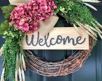 May include: A grapevine wreath with a burlap bow and pink hydrangea flowers. A wooden sign with the word "Welcome" is attached to the wreath.