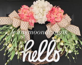 May include: A spring wreath with pink and white hydrangeas, greenery, and a burlap bow. The wreath has a white wooden sign that says "hello".