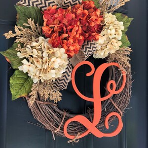 May include: A fall-themed wreath with a burlap bow, orange and white hydrangeas, and a large orange wooden letter "L".