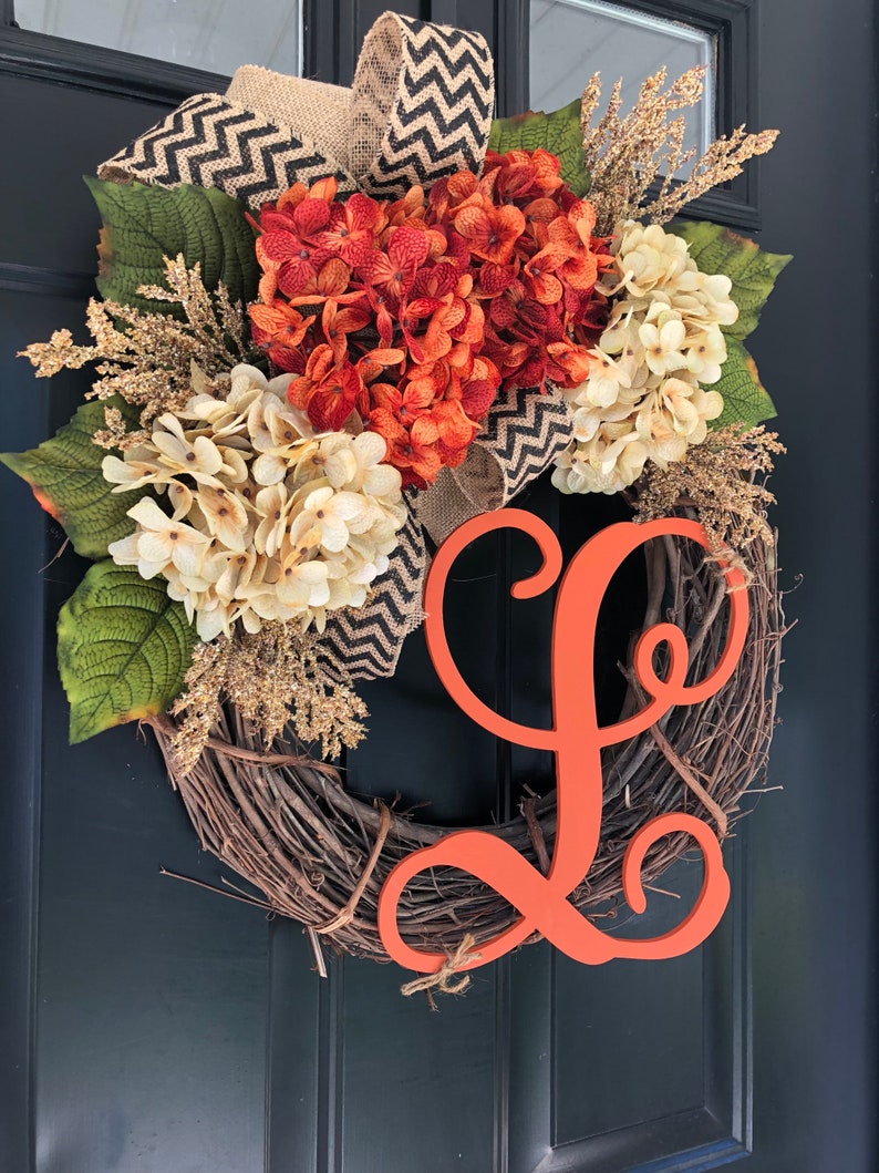 May include: A fall-themed wreath with a burlap bow, orange and white hydrangeas, and a large orange wooden letter "L".