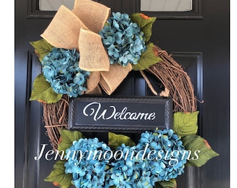 May include: A blue and green wreath with a burlap bow and a black sign that says "Welcome".