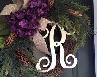 May include: A wreath with a large white letter "R" in the center. The wreath is made of pine branches and has purple hydrangeas, gold ribbon with white polka dots, and pine cones.