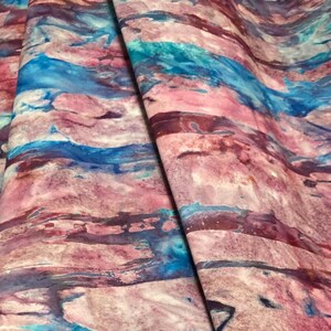 May include: Fabric with a watercolor-like design in shades of pink, blue, and purple. The fabric has a marbled appearance with flowing, abstract patterns. The colors blend together, creating a visually appealing textile.