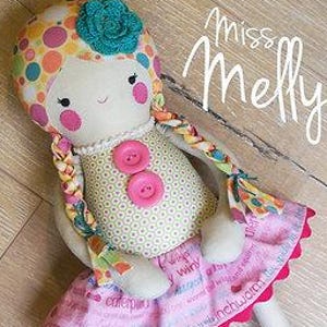 May include: A handmade fabric doll with a polka-dot head, braided hair, and a floral hat. The doll has a yellow patterned top with pink buttons and a pink skirt with a scalloped edge. The doll's legs are white with blue boots. The text "Miss Melly" is written on the image.