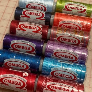 May include: A collection of colorful Omega brand nylon thread spools. The spools are in various colors, including blue, red, green, and purple. Each spool has a label with the Omega logo and text indicating it is 100% nylon thread.