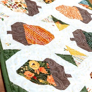 May include: A quilted table runner featuring a repeating pattern of acorns and triangles. The acorns have brown caps and colorful patterned bodies in shades of orange, yellow, and floral prints. The background is white with small yellow dots and green trim.