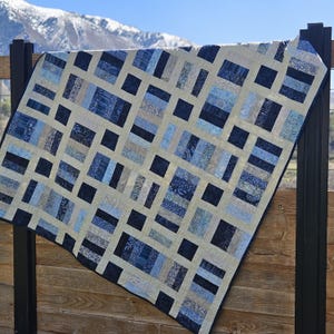 May include: A blue and cream patchwork quilt with a grid pattern. The quilt is made of various shades of blue and cream fabric, and the grid pattern is created by using different widths of fabric.