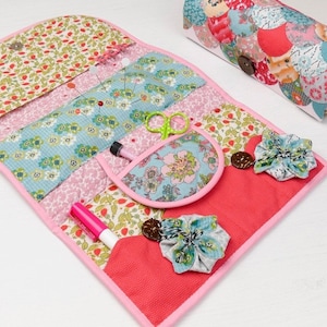 May include: A pink fabric sewing kit with multiple pockets and compartments. The kit is open and shows various sewing supplies, including pins, scissors, a marker, and a small fabric flower.