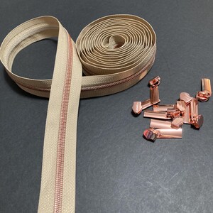 May include: A beige zipper with a copper-colored zipper pull and teeth, partially rolled up. Several copper-colored zipper end pieces are scattered nearby. The zipper is on a dark gray surface.