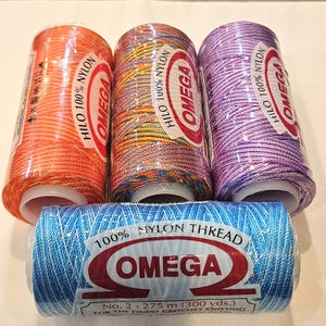 May include: Four spools of Omega Hilo 100% Nylon thread in various colors. The colors include orange, a mix of rainbow hues, purple, and blue. The blue spool has the text "100% Nylon Thread" and "Omega". The thread is for crochet knitting.