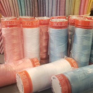 May include: A close-up of a variety of spools of thread in different colors, including pink, white, blue, and peach. The spools are labeled "100% Cotton" and "Aurifil".