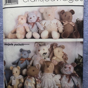May include: A Simplicity sewing pattern for making teddy bears. The pattern is for one size and features a variety of teddy bear designs, including a bear wearing a dress, a bear wearing a bow tie, and a bear wearing a floral print. The pattern is titled "crafts/ouvrages" and is designed by Marjorie Puckett.