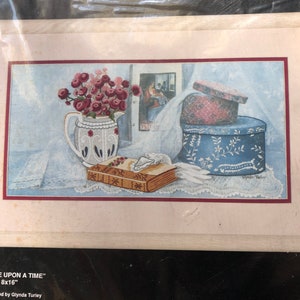 May include: A framed watercolor painting titled "Once Upon a Time" featuring a still life. The artwork depicts a white pitcher with red flowers, a book, a blue hat box, and a framed picture. The painting is 8x16 inches.