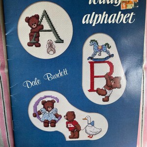 May include: A blue softcover book titled Teddy's Alphabet by Dale Burdett, published by Burdett Publications. The cover features cross-stitch illustrations of teddy bears with the letters A, B, and C, alongside a rocking horse and a duck.