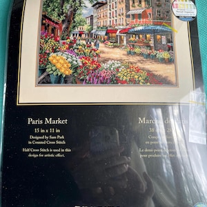 May include: A Dimensions The Gold Collection counted cross stitch kit titled Paris Market, designed by Sam Park. The packaging features a 15 in x 11 in finished design of a colorful flower market on a street in Paris with buildings and trees.