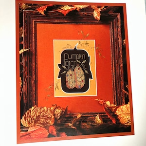 May include: A framed cross stitch design featuring a black pumpkin with the text "Pumpkin Patch" in white. The frame is surrounded by fall leaves in shades of orange, brown, and yellow.