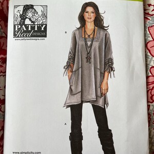 May include: A Simplicity 1543A sewing pattern envelope featuring Patty Reed Designs. The illustration shows a grey tunic with a square neckline, side pockets, and gathered sleeves with ties, paired with black leggings and black boots.