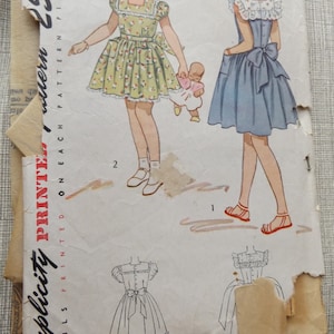 Może przedstawiać: Vintage wzór Simplicity na sukienki dla dziewczynek, rozmiar 8. Ilustracja przedstawia dwie dziewczynki w sukienkach, jedna w żółte kwiaty, druga w niebieską. Szczegóły wzoru są wydrukowane na przodzie, w cenie 25 centów.