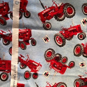 May include: Light blue fabric featuring a repeating pattern of red Farmall tractors and white International Harvester logos. A white selvage edge runs vertically along the left side with the text 2017 CNH Industrial America printed on it.