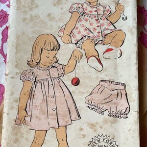 May include: Vintage sewing pattern for a child's dress and bloomers. The pattern is for size 1 and is from New York Pattern. The pattern is illustrated with two girls wearing the dress and bloomers. The pattern is in pink and white.