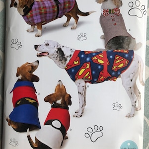 May include: Five different dog clothing patterns featuring various superhero themes. The patterns include a plaid design, a Superman design, a Wonder Woman design, a Pokemon design, and a tuxedo design. The patterns are printed on a white background with paw prints.
