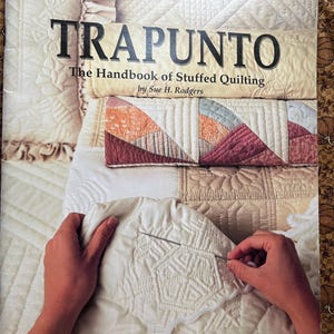 May include: A softcover instructional book titled Trapunto: The Handbook of Stuffed Quilting by Sue H. Rodgers. The cover features a photograph of hands using a needle and white yarn to create stuffed quilting on a cream fabric surface.