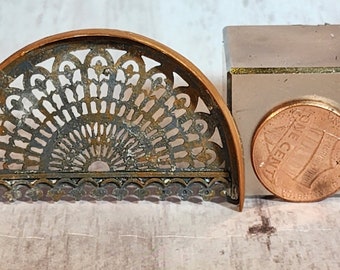 May include: A decorative, half-circle metal piece with an intricate, openwork design. The piece is copper-coloured and has a detailed, fan-like pattern. Next to it is a small, square, light-coloured object and a penny.