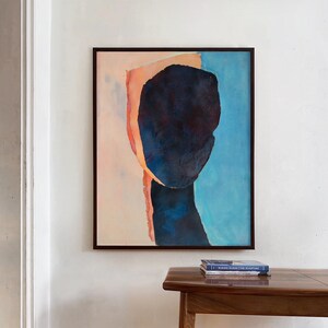A wall decor with an art print of an abstract portrait in peach fuzz, shades of dark indigo blue on a pale salmon and sky blue background. The work is composed of three torn paper layers with rough edges and wrinkled texture in a black minimal frame.