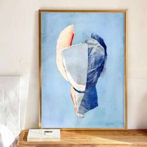 May include: Abstract art print in a gold frame. The artwork features a collage of torn paper in shades of blue, white, and red against a light blue background. The artwork is displayed on a wooden surface.