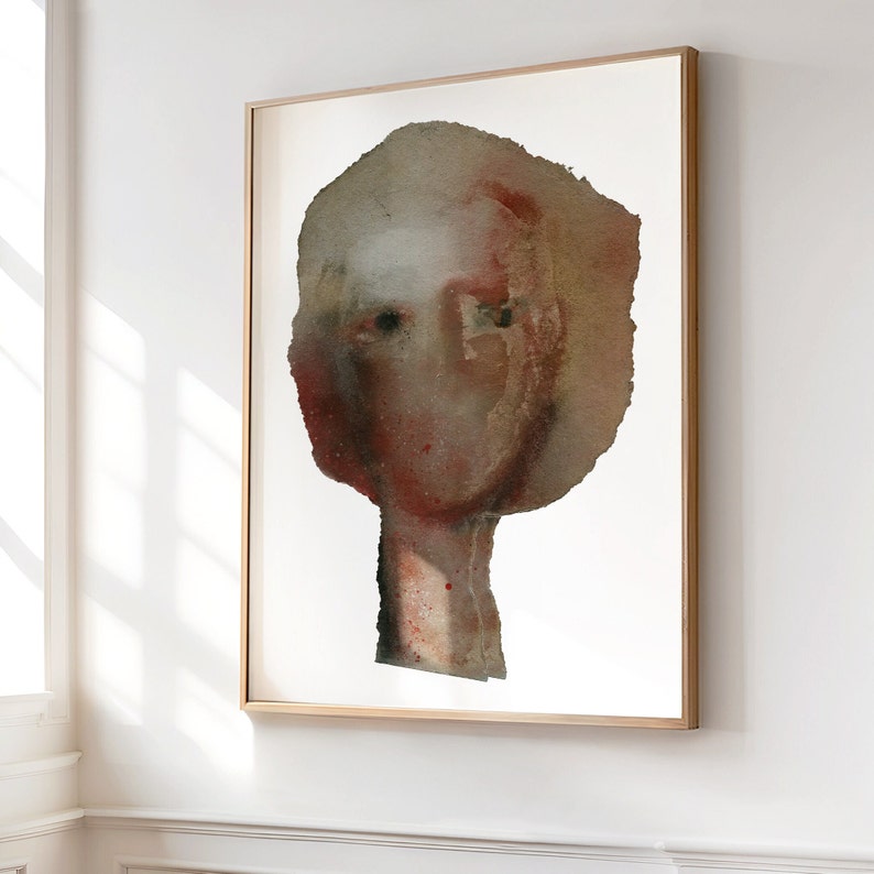 A framed high quality art print of a modern watercolor female portrait in maroon, beige, brown and grey. Two spray painted thick paper layers are torn to shape, with rough edges. A vague nose, scratched off the paint, hair line and two black eyes.