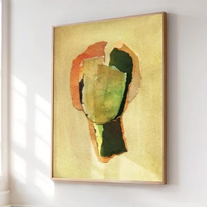 May include: Abstract art print in a wooden frame. The artwork features layered paper in shades of green, orange, and yellow against a light yellow background. The frame is a light brown color. The artwork is displayed on a white wall.