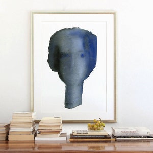 Puede incluir: Una pintura al agua enmarcada de un rostro abstracto azul y gris. La pintura est&aacute; colgada en una pared blanca sobre una pila de libros y un cuenco de uvas verdes.