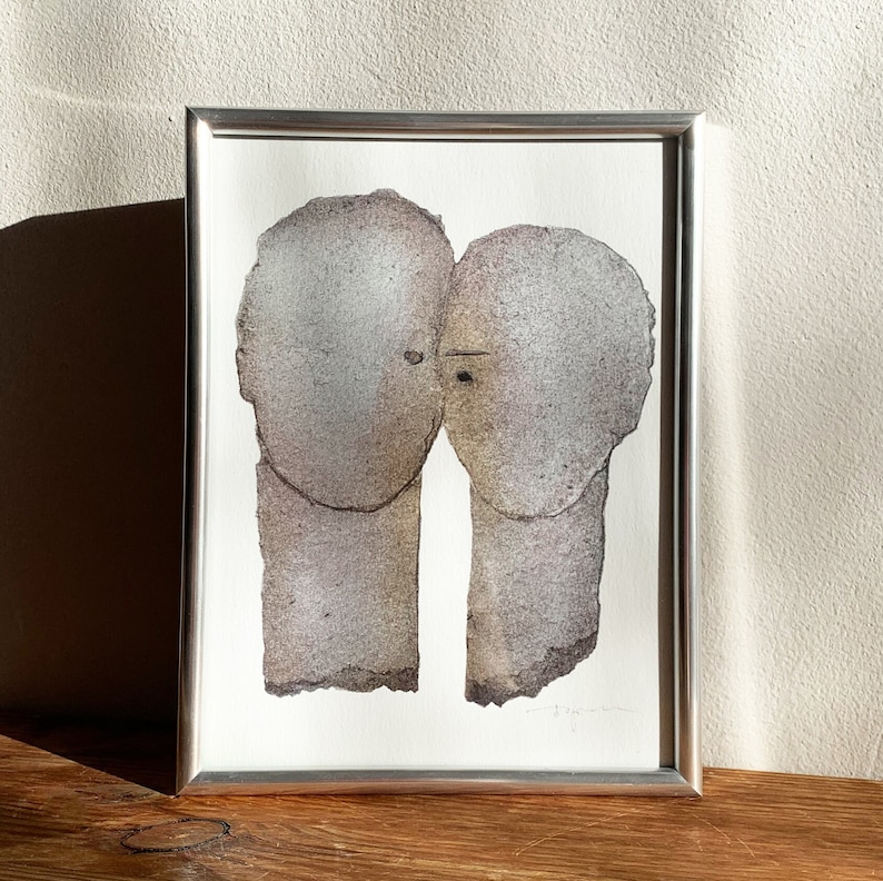 A decor of a silver framed art print of a romantic abstract portrait of a love couple in shades of grey with silver finish on old wood. The two heads are composed of torn rough paper layers with exposed grungy edges and two eyes for accent.