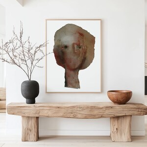 A light framed high quality art print of a modern watercolor-effect portrait in maroon, beige, brown, grey. Two spray painted thick paper layers are torn to shape, with rough edges. A vague nose, scratched off the paint, hair line and two black eyes.