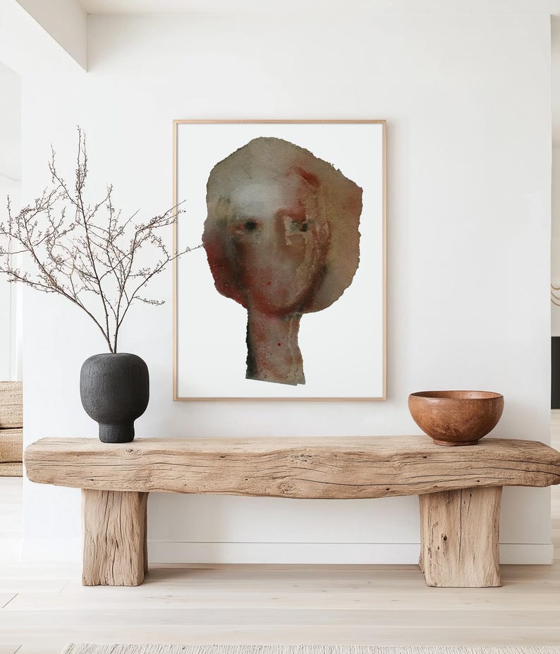 A light framed high quality art print of a modern watercolor-effect portrait in maroon, beige, brown, grey. Two spray painted thick paper layers are torn to shape, with rough edges. A vague nose, scratched off the paint, hair line and two black eyes.