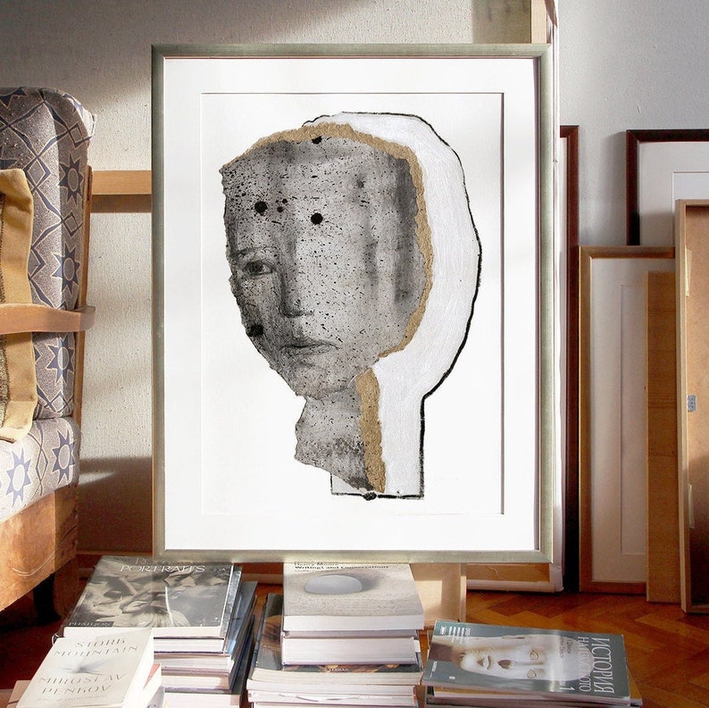 A living room decor of a golden framed high quality art print of a modern abstract portrait in shades of gray. A layer of ripped thick paper has ochre rough edges and graphite sketched facial features. A bigger head shape is outlined with charcoal.