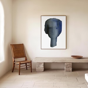 Puede incluir: Una pintura al agua enmarcada de un rostro abstracto azul y gris sobre un fondo blanco. La pintura est&aacute; colgada en una pared blanca sobre un banco de piedra largo de color gris claro.
