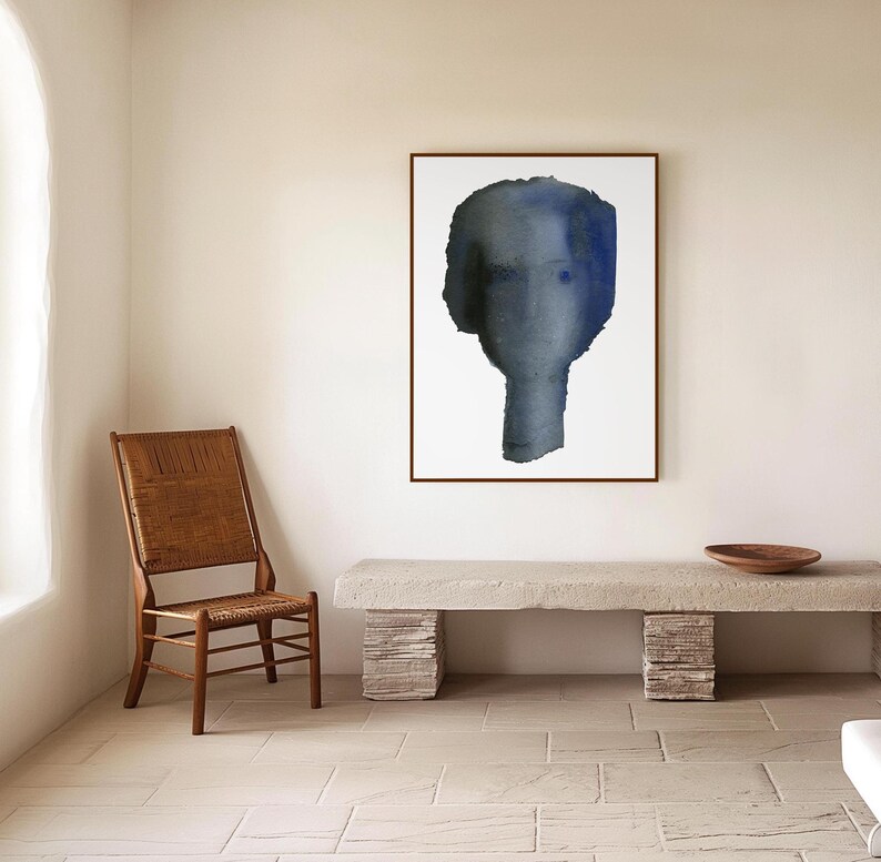 Puede incluir: Una pintura al agua enmarcada de un rostro abstracto azul y gris sobre un fondo blanco. La pintura est&aacute; colgada en una pared blanca sobre un banco de piedra largo de color gris claro.
