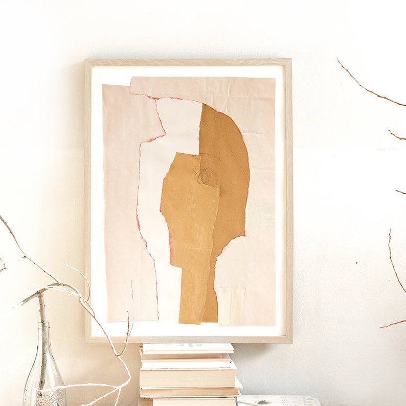 A cosy room decor with a fine quality art print of an abstract portrait. It is in a tender peach yellow pallet, with torn paper edges in cherry red, a pencil drawn eye for an accent. Layers in desert sand cut out a head from a pale peach background.