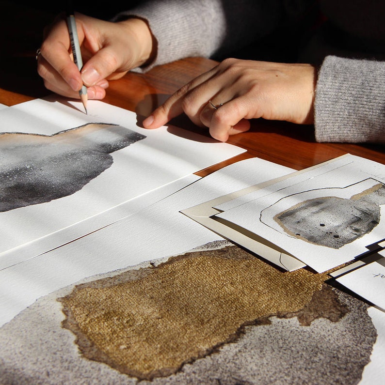 An artist hand signs a grey fine surface minimal art print of an original abstract portrait, printed with high fade resistance inks, It is on a lacquered wood table with another, gold and grey art print of another portrait, a postcard, an envelope.