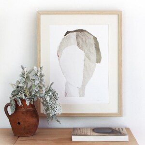 A decor with a clay pot with greens and a light framed art print of an original abstract portrait in greige, of-white and ecru on a pile of art books. The edges of the thick paper torn layers outline the head on white for a minimal graphic feeling.