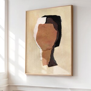 May include: Abstract art print featuring a collage of torn paper pieces in shades of brown, black, and white, forming a human face. The artwork is framed in a light wood frame and set against a beige background.