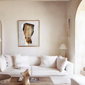May include: A white sofa with white cushions in a living room with a large abstract painting on the wall. The painting is in a gold frame and features a brown and white abstract design. There is a wooden coffee table in front of the sofa with a decorative sculpture on it.