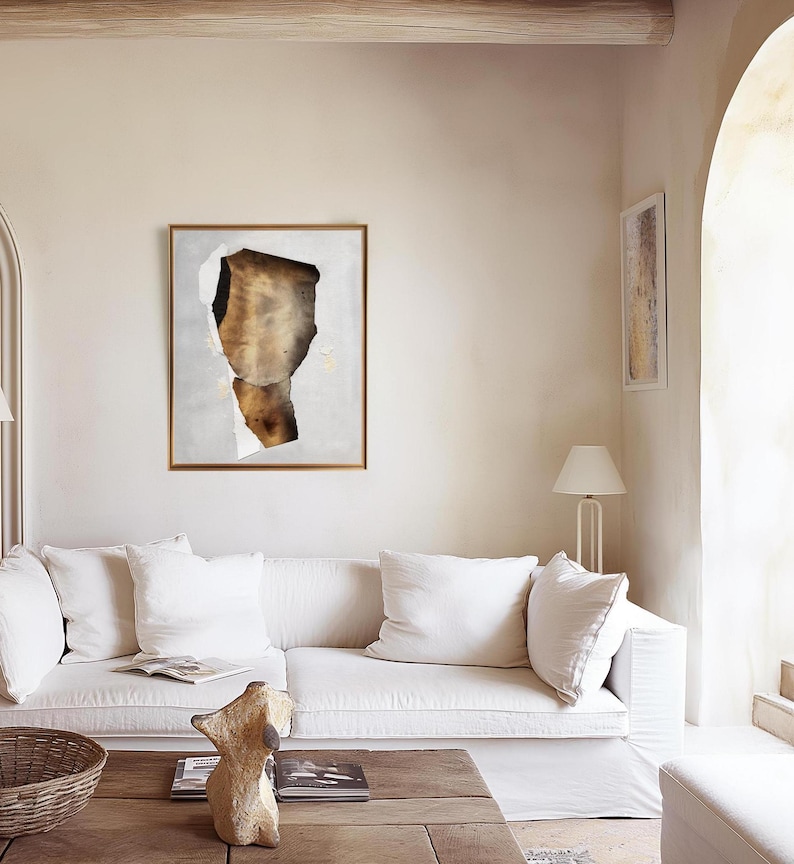 May include: A white sofa with white cushions in a living room with a large abstract painting on the wall. The painting is in a gold frame and features a brown and white abstract design. There is a wooden coffee table in front of the sofa with a decorative sculpture on it.